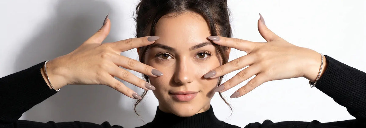 Short almond press-on nails in a soft greige neutral from the de blinkie X Collection, worn on both hands framing the face.
