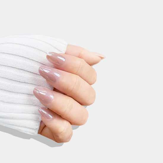 Closed hand pose showing de blinkie Barely Mocha rosy mocha nude press-on nails in short oval shape against light grey background with white knit sleeve