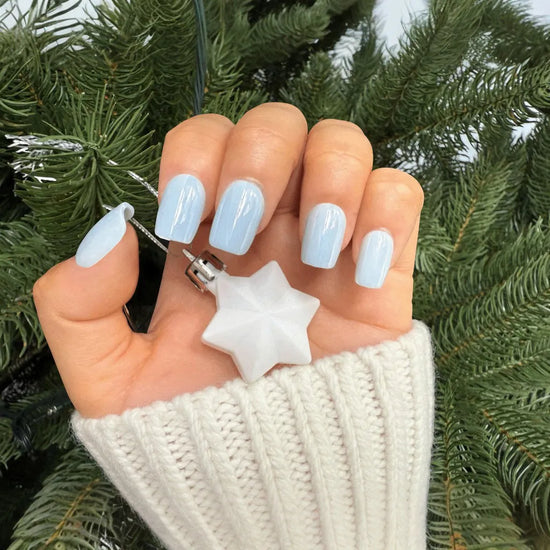 Closed fist holding white star Christmas ornament showing de blinkie Bluebell Dream baby blue short square press-on nails against festive pine tree background