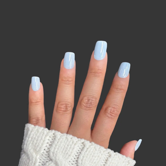 Upright spread hand showing Bluebell Dream clean baby blue soft gloss short square press-on nails against dark grey background with white knit sleeve