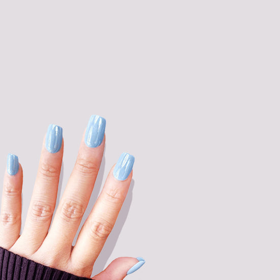 Folded fingers showing de blinkie Bluebell Glaze soft baby blue glazed press-on nails in short square against a light grey background with a charcoal ribbed knit sleeve