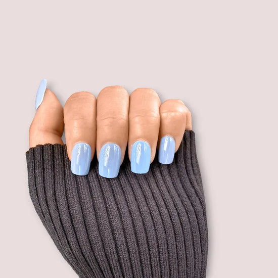 Claw pose showing de blinkie Bluebell Glaze soft baby blue glazed press-on nails in short square against a soft blush grey background with a charcoal ribbed knit sleeve