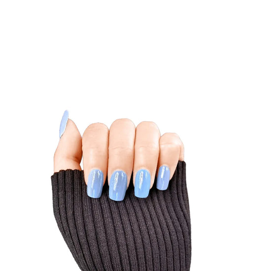 Claw pose showing de blinkie Bluebell Glaze soft baby blue glazed press-on nails in short square against a white background with a charcoal ribbed knit sleeve