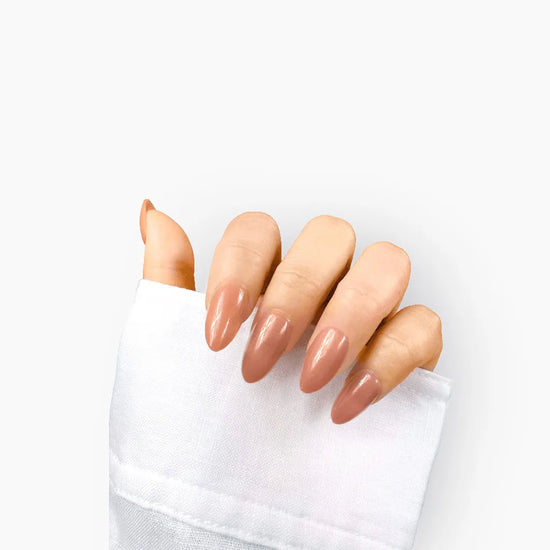 Claw pose showing de blinkie Blush Apricot warm peachy nude press-on nails in short almond shape against white background with white linen sleeve