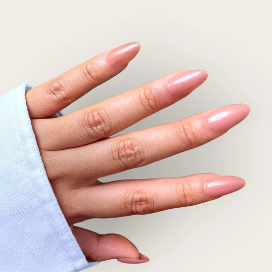 Horizontal spread hand showing de blinkie Blush Apricot warm peachy nude press-on nails in short almond shape against warm cream background with light blue sleeve