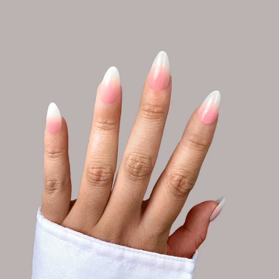 Upright hand pose showing de blinkie Blush Elise pink to white ombre press-on nails in short almond shape against grey background