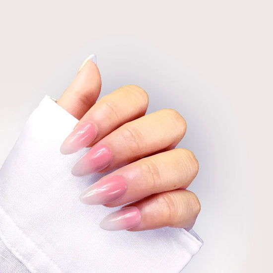 Close-up of de blinkie Blush Elise glazed pink to white gradient press-on nails in short almond shape, closed hand pose with white sleeve