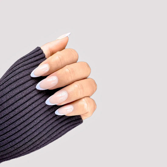 Claw pose showing de blinkie Blush Mirage warm nude and aurora iridescent French tip press-on nails in short almond against a light grey background with a charcoal ribbed knit sleeve