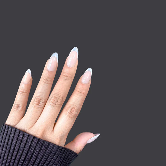 Fingers spread upward showing de blinkie Blush Mirage warm nude and aurora iridescent French tip press-on nails in short almond against a dark charcoal background with a black ribbed knit sleeve