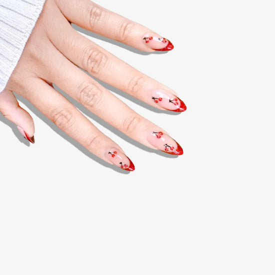 Downward hand pose showing de blinkie Cherry Blush sheer nude with cherry 3D detail and red glitter tips press-on nails in short almond shape against grey background with white ribbed knit sleeve