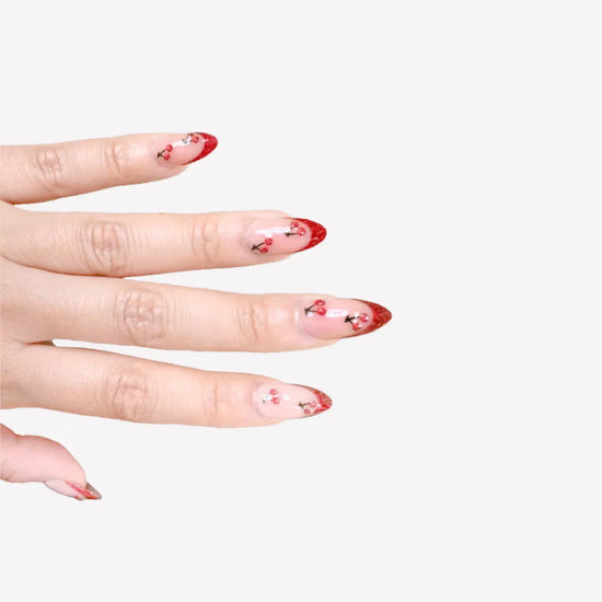 Fingers spread pose showing de blinkie Cherry Blush sheer nude with cherry 3D detail and red glitter tips press-on nails in short almond shape against grey background