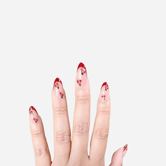 Upward hand pose showing de blinkie Cherry Blush sheer nude with cherry 3D detail and red glitter tips press-on nails in short almond shape against grey background