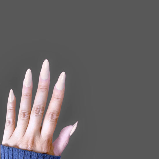 Diagonal downward hand showing de blinkie Creme Glazed sheer milky cream glazed press-on nails in medium almond against a dark grey background with a navy ribbed knit sleeve