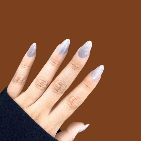 Diagonal hand pose showing de blinkie Crystal Muse frosted icy silver shimmer short almond press-on nails against brown background