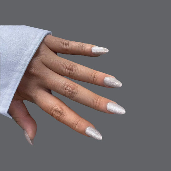 Downward hand pose showing de blinkie Crystal Muse icy silver shimmer short almond press-on nails with white sleeve against grey background