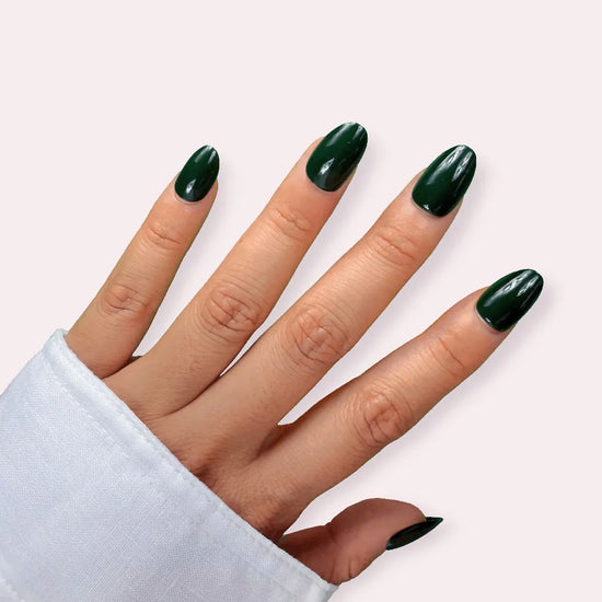 Diagonal spread hand showing de blinkie Emerald Envy deep jewel-toned forest green press-on nails in short oval shape against pale pink background with white sleeve