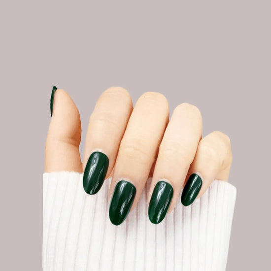 Upright closed hand pose showing de blinkie Emerald Envy deep jewel-toned forest green press-on nails in short oval shape against warm mauve background with white ribbed sleeve