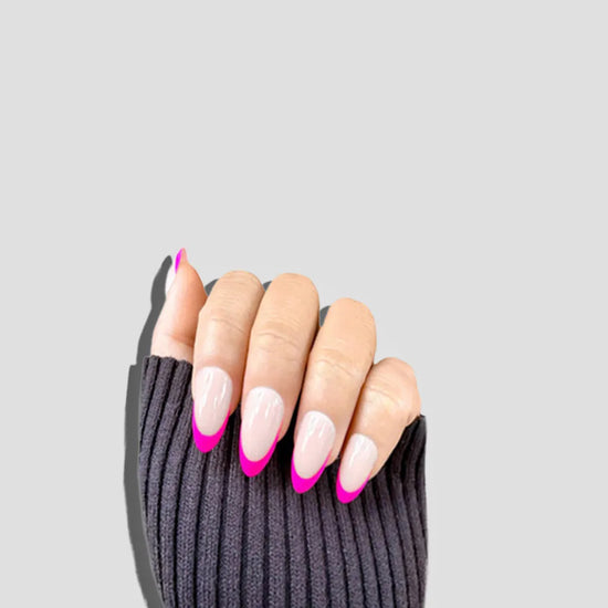 Claw pose showing de blinkie Fleur Fuchsia sheer nude base with vivid fuchsia curved French tip press-on nails in short almond against a soft light grey background with a dark charcoal ribbed knit sleeve