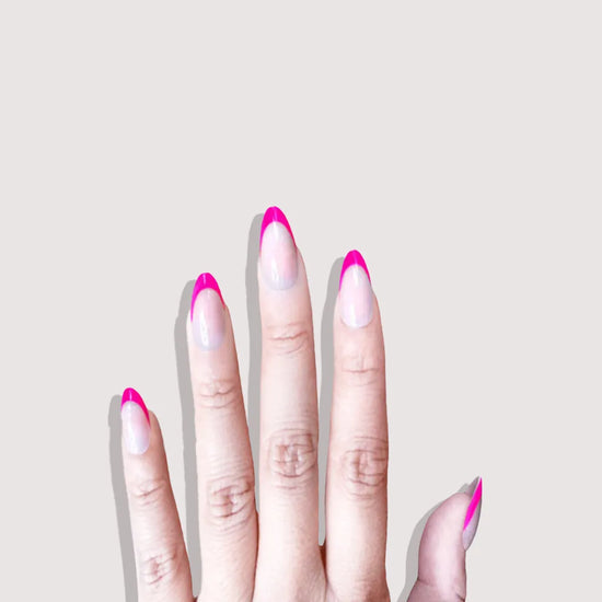 Downward spread hand showing de blinkie Fleur Fuchsia sheer nude base with vivid fuchsia curved French tip press-on nails in short almond against a soft light grey background with a dark charcoal ribbed knit sleeve