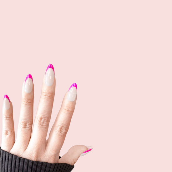 Spread hand showing de blinkie Fleur Fuchsia sheer nude base with vivid fuchsia curved French tip press-on nails in short almond against a soft pink background with a dark charcoal ribbed knit sleeve