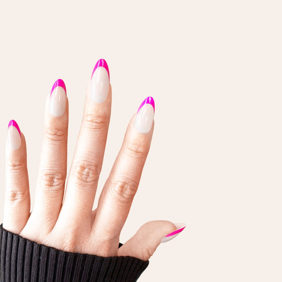 Spread hand showing de blinkie Fleur Fuchsia sheer nude base with vivid fuchsia curved French tip press-on nails in short almond against a warm cream background with a dark charcoal ribbed knit sleeve
