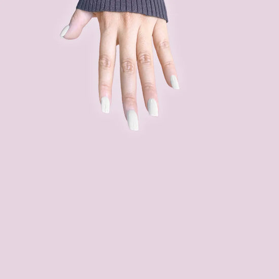 Fingers pointing downward showing de blinkie Glazed Milkyway milk white gloss press-on nails in short square against a soft lilac background with a charcoal ribbed knit sleeve