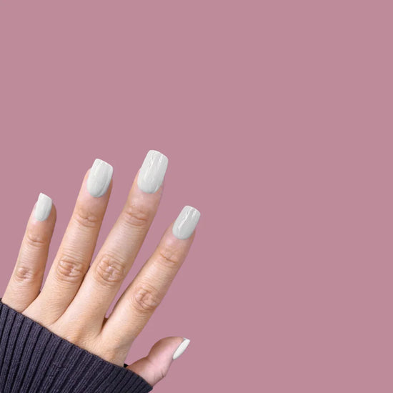 Fingers spread upward showing de blinkie Glazed Milkyway milk white gloss press-on nails in short square against a warm mauve background with a charcoal ribbed knit sleeve