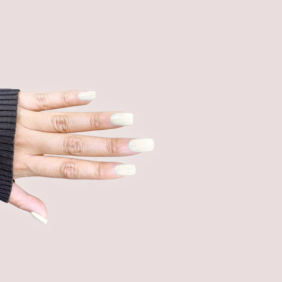 Spread hand showing de blinkie Glazed Milkyway milk white gloss press-on nails in short square against a soft blush pink background with a charcoal ribbed knit sleeve