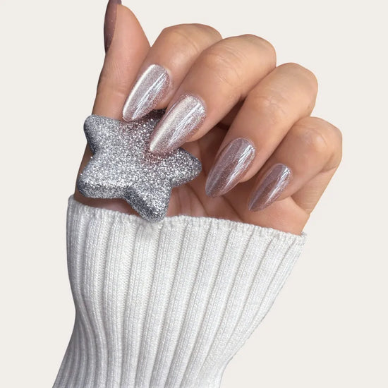 Claw hand holding silver glitter star ornament showing de blinkie Glazed Mocha rosy mauve velvet cat eye short almond press-on nails against cream background with white knit sleeve