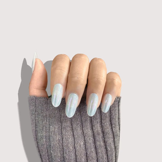 Claw pose showing de blinkie Glazed Stardust sheer pearl white opalescent shimmer press-on nails in medium oval shape against pale grey background with dark grey ribbed sleeve