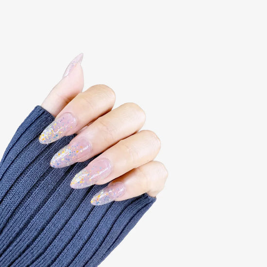 Claw pose showing de blinkie Glitter Drip sheer pink nude base with chunky multicolour glitter press-on nails in medium almond against a soft light grey background with a dark navy ribbed knit sleeve