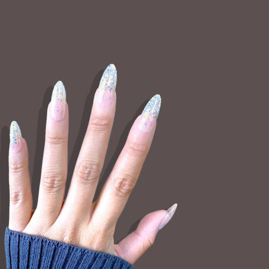 Upward spread hand showing de blinkie Glitter Drip sheer pink nude base with chunky multicolour glitter press-on nails in medium almond against a dark brown background with a dark navy ribbed knit sleeve