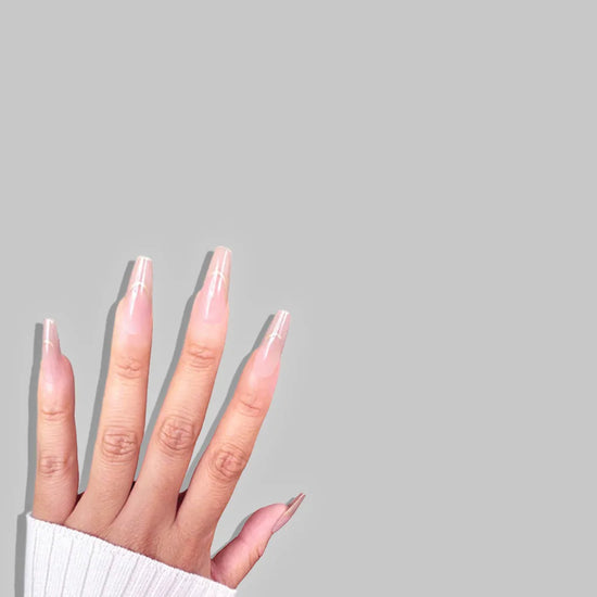 Fingers pointing upward showing de blinkie Golden Grace sheer pink nude with gold trace press-on nails in long coffin against a soft grey background with a white ribbed knit sleeve