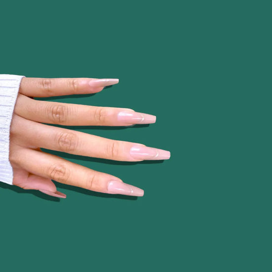 Side-angled hand showing de blinkie Golden Grace sheer pink nude with gold trace press-on nails in long coffin against a deep forest green background with a white ribbed knit sleeve
