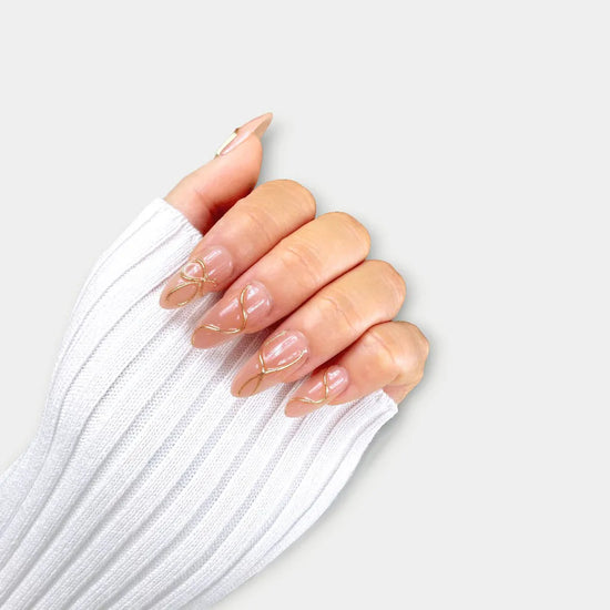 Claw pose showing de blinkie Golden Trace golden nude with 3D gold swirl detail press-on nails in medium almond shape against grey background with white ribbed knit sleeve
