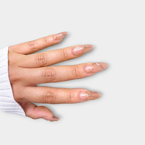 Diagonal hand pose showing de blinkie Golden Trace golden nude with 3D gold swirl detail press-on nails in medium almond shape against grey background with white ribbed knit sleeve