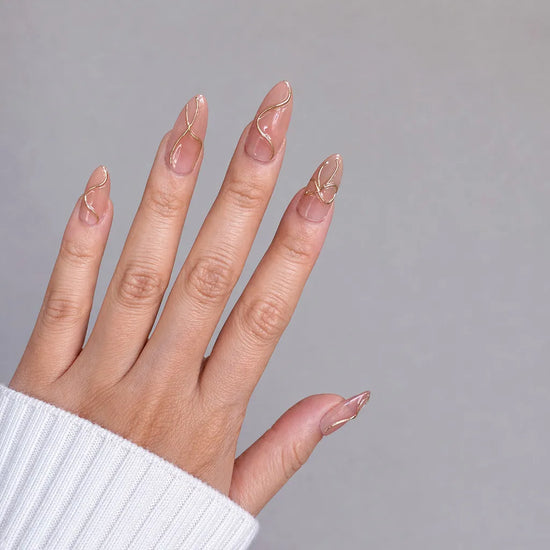 Fingers spread pose showing de blinkie Golden Trace golden nude with 3D gold swirl detail press-on nails in medium almond shape against grey background with white ribbed knit sleeve