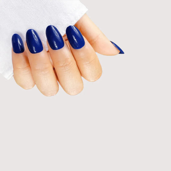 Claw pose showing de blinkie Indigo Ivy rich indigo blue press-on nails in short oval shape against light grey background with white linen sleeve