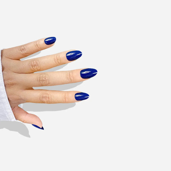Diagonal hand pose showing de blinkie Indigo Ivy rich indigo blue press-on nails in short oval shape against light grey background with white sleeve