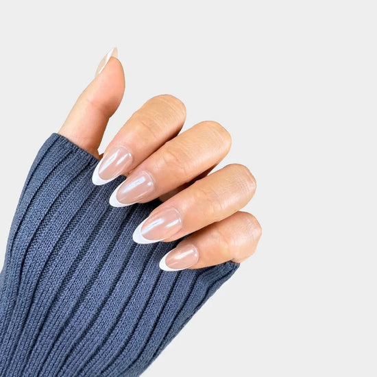 Claw pose showing de blinkie Ivory Wave sheer nude and white French tip press-on nails in short almond against a light grey background with a denim blue ribbed knit sleeve