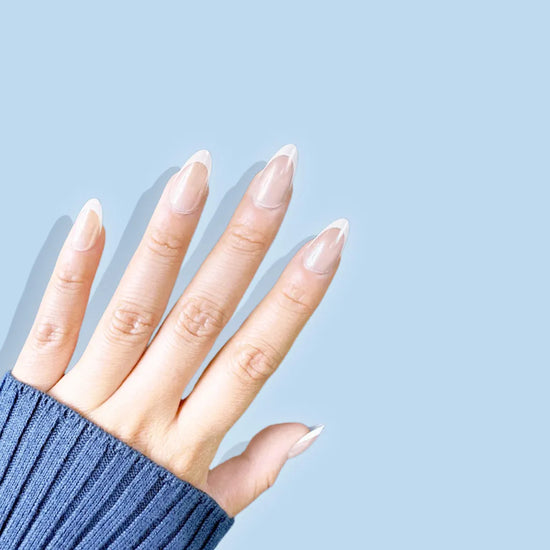 Fingers spread upward showing de blinkie Ivory Wave sheer nude and white French tip press-on nails in short almond against a sky blue background with a denim blue ribbed knit sleeve