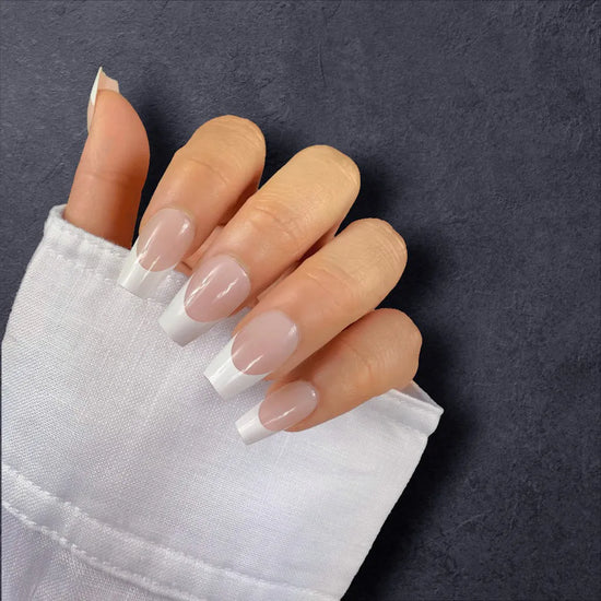 Claw pose showing de blinkie La Belle Frenchie sheer blush pink French tip press-on nails in short coffin shape against dark charcoal textured background with white linen sleeve