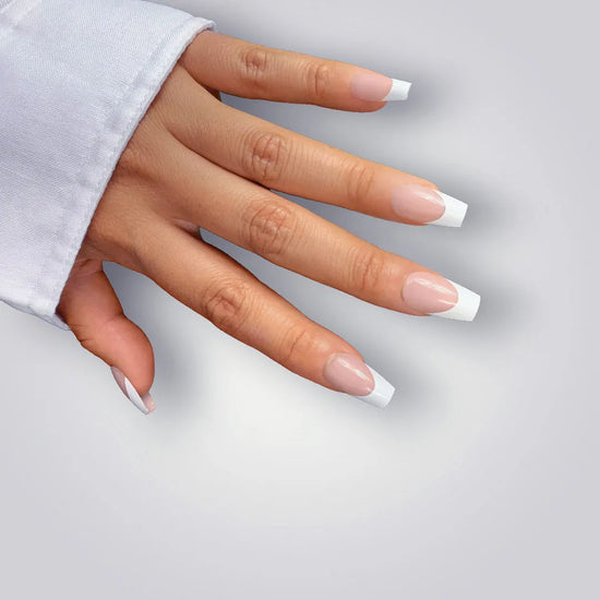 Down-facing hand pose showing de blinkie La Belle Frenchie sheer blush pink French tip press-on nails in short coffin shape against light grey background with white sleeve