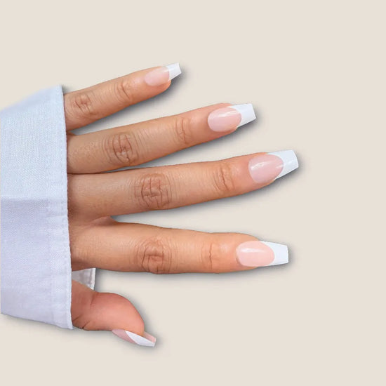 Spread hand showing de blinkie La Belle Frenchie sheer blush pink French tip press-on nails in short coffin shape against cream background with white linen sleeve