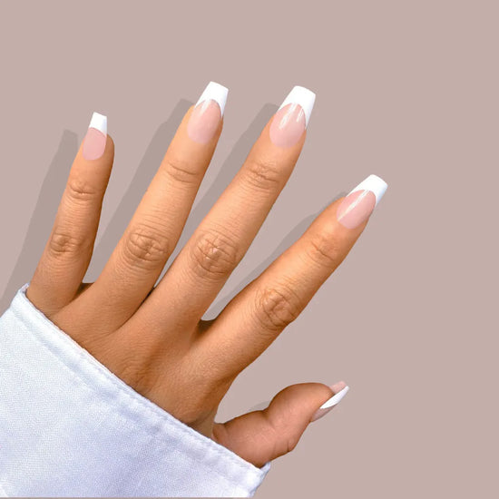 Spread hand showing de blinkie La Belle Frenchie sheer blush pink French tip press-on nails in short coffin shape against warm mauve background with white sleeve