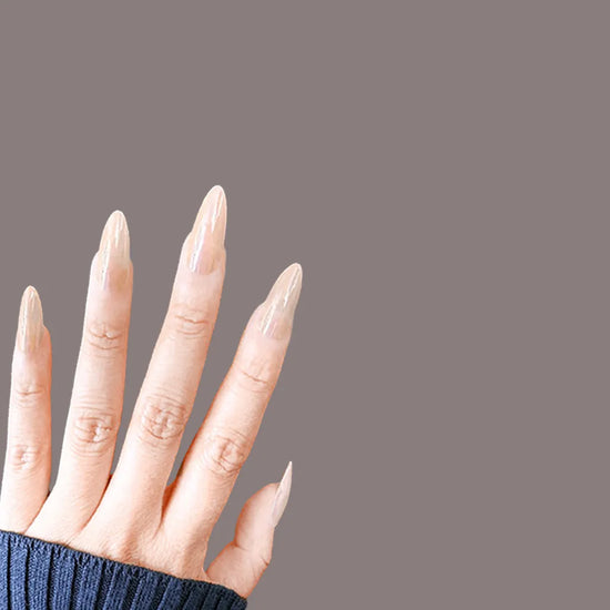 Fingers-spread hand angled left-downward showing de blinkie Lady Aurora sheer nude press-on nails in medium almond against a warm taupe background with a navy ribbed knit sleeve