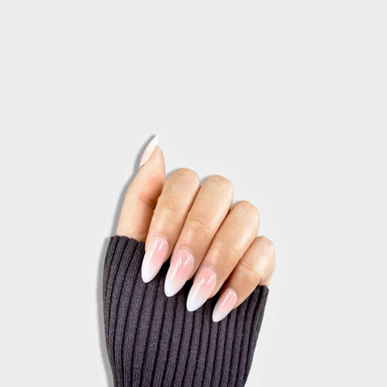 Claw pose showing de blinkie Lady Haze blush pink to white ombre press-on nails in short almond against a light grey background with a dark charcoal ribbed knit sleeve