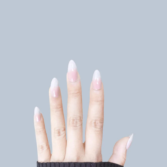 Diagonal left-facing hand showing de blinkie Lady Haze blush pink to white ombre press-on nails in short almond against a soft grey-blue background with a black knit sleeve