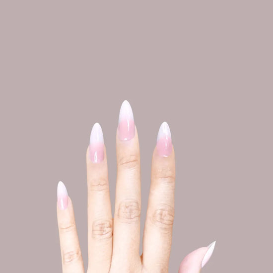 Downward-facing hand showing de blinkie Lady Haze blush pink to white ombre press-on nails in short almond against a warm taupe background with no sleeve visible