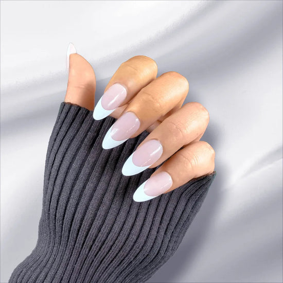 Claw pose showing de blinkie Lady Primrose soft sheer pink base with bold white French tip press-on nails in medium almond shape against light grey draped fabric background with dark charcoal ribbed sleeve